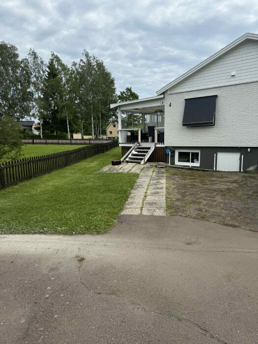 Hus med vit fasad, veranda, trappa och gräsmatta med ny klippyta för robotgräsklippare. Staket och träd i bakgrunden.