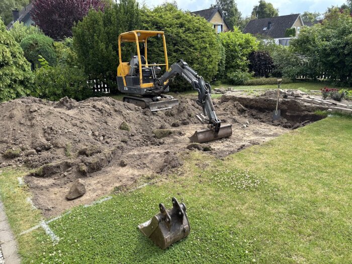 Minigrävare vid en uppgrävd tomt och jordhög; förberedelse för att bygga en 15m2 friggebod som ska fungera som verkstad.