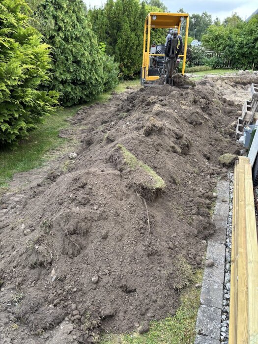 Minigrävare och stor jordhög vid sidan av en trädgård, omgiven av buskar och träd. Förbereder grund för friggebod/verkstad.