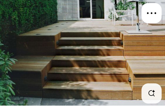 Träterrass med trappsteg, perfekt för sittplatser. Vänster sida har bredare ytor nära en tät häck, skuggor skapar mönster över trappan.