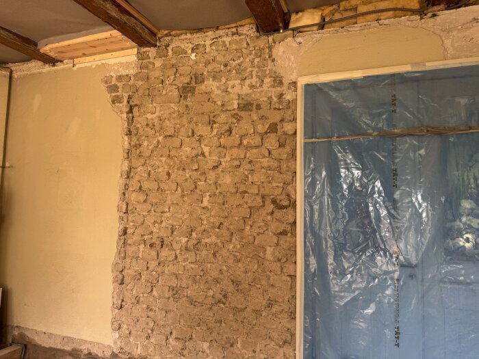 Grov tegelvägg delvis avtäckt till vänster, plastskydd höger, visar renovering av gammal yttervägg i lertegel.