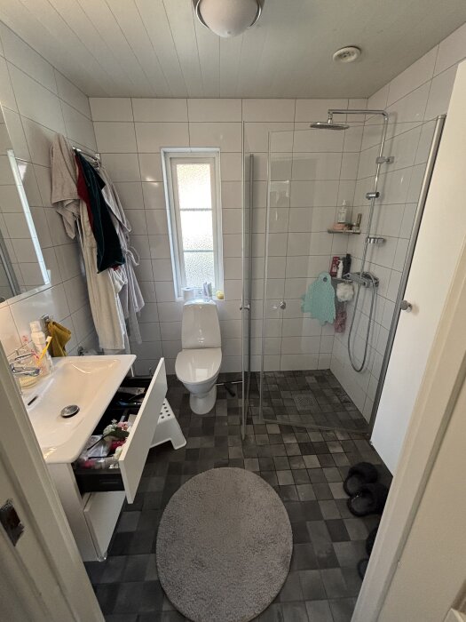 Badrum med duschkabin och handfat. Öppen låda under handfatet. Möjligt läckage vid rören där de ansluter till väggen. Duschhuvud och toalett synliga.