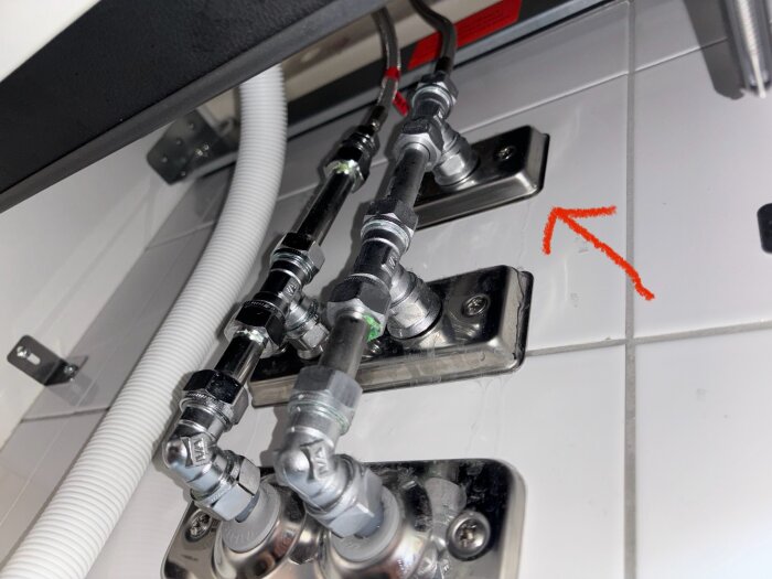 Röranslutningar under ett handfat, med en röd pil som markerar ett läckage från väggen. Vit kakel bakgrund.