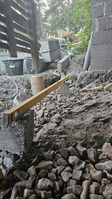 Murade väggar av lättklinkerblock under en terass, med grus på marken och ett vattenpass för att jämna ut nivåskillnader.