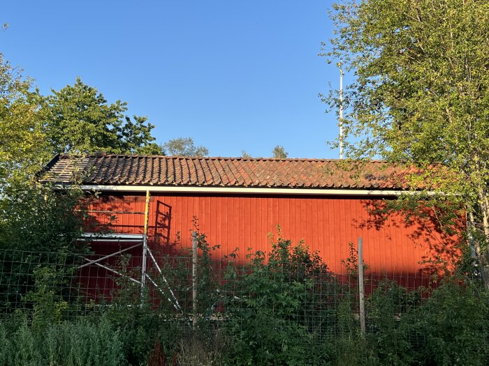 En röd byggnad med tak av modifierade tegeltakpannor, omgiven av grönska och ett trästaket.