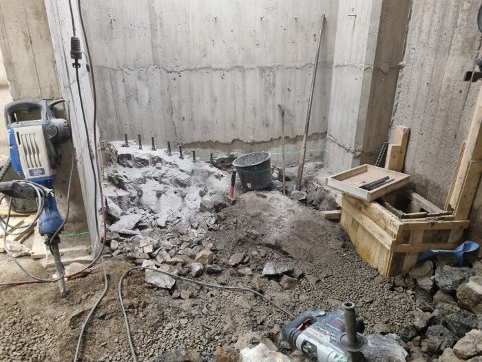 Byggarbetsplats med borrmaskin, betongväggar och verktyg, omgiven av sten och jord, förbereder för vidare konstruktion.