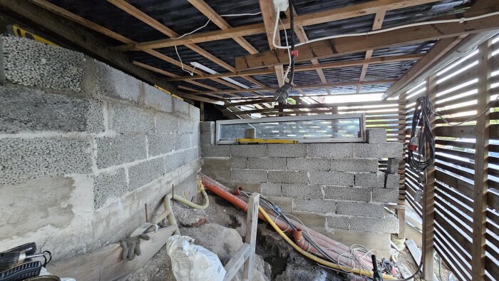 Öppet förråd under terrass, väggar i lättklinkerblock, synliga rör och sladdar, del av betonggolv synligt, pågående renovering.
