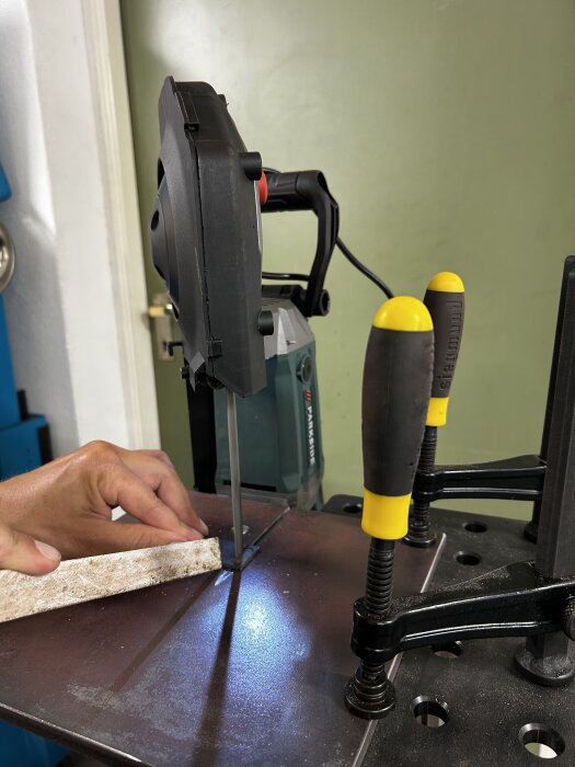 Modifierad bandsåg med hand som guidar ett trästycke genom sågbladet, verktyg med gult handtag syns i bakgrunden.