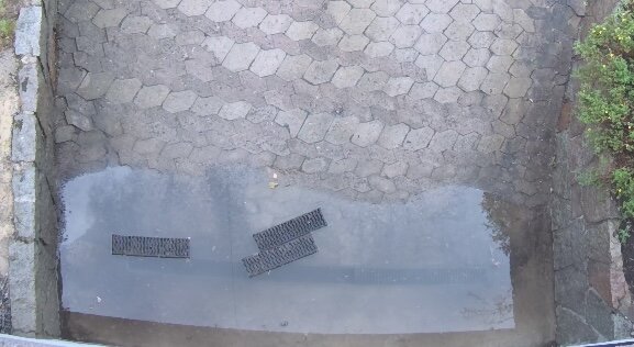 Vattenpöl på stenlagd mark vid garageinfart, med två dräneringsgaller delvis täckta av vatten.