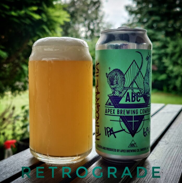 En ölglas fyllt med oklar gul vätska bredvid en burk med etiketten "Apex Brewing Company IPA" på ett bord utomhus.