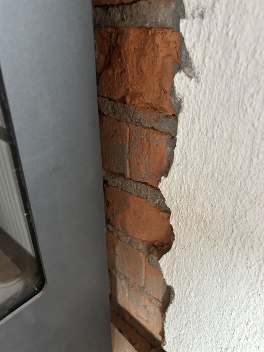 Råtegelvägg med skorstensmur vid sidan av en plåtbeklädd kamin, intill grov vitmurad yta som behöver återställas.