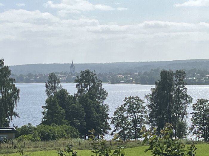 Sjölandskap med gröna träd i förgrunden, en kyrka och husdjur mellan trädtopparna på andra sidan vattnet under en delvis molnig himmel.