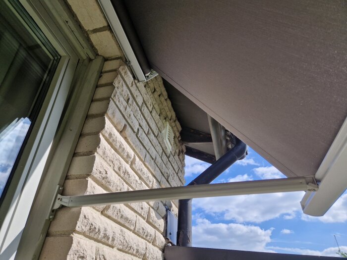 Markis ovanför ett fönster på en tegelvägg, med del av avloppsrör och klar himmel i bakgrunden.