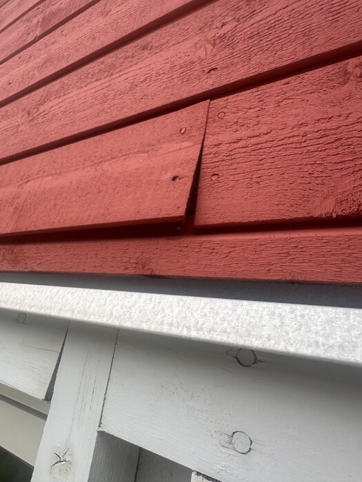 Röd träpanel med en planka som sticker ut på en fasad, ovanför en vit metallkant.