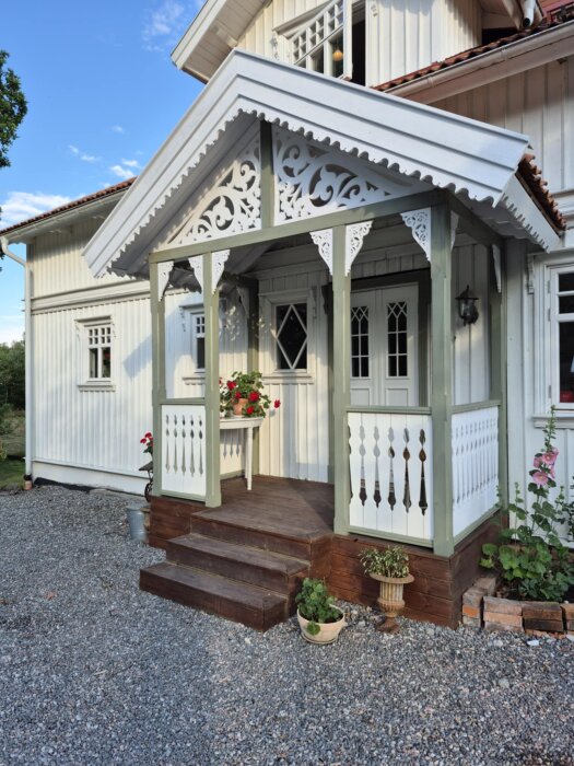 Dekorerad och färdigställd farstukvist med pelare, handledare och vindskivor, omgiven av grus och planterade blommor framför ett ljusmålat hus.