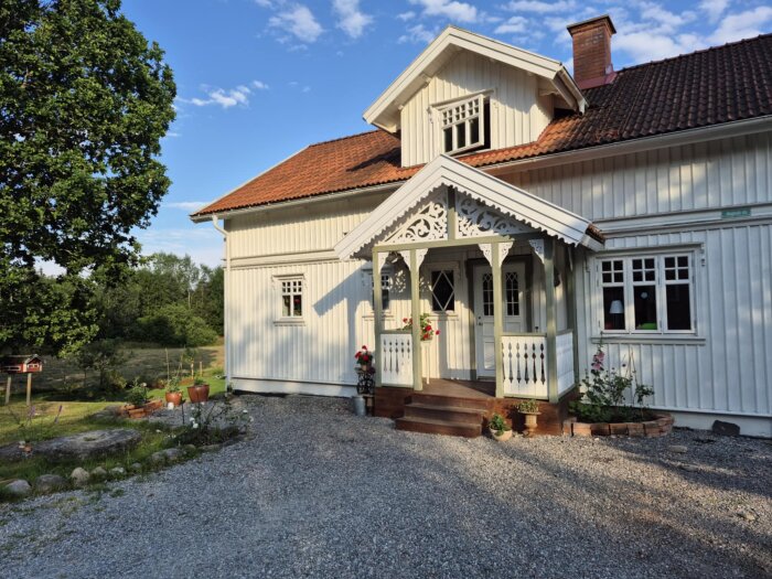 Farstukvist på vit trävilla med detaljrika snickerier och blommor; hus med rött tegeltak och trädgård i bakgrunden.