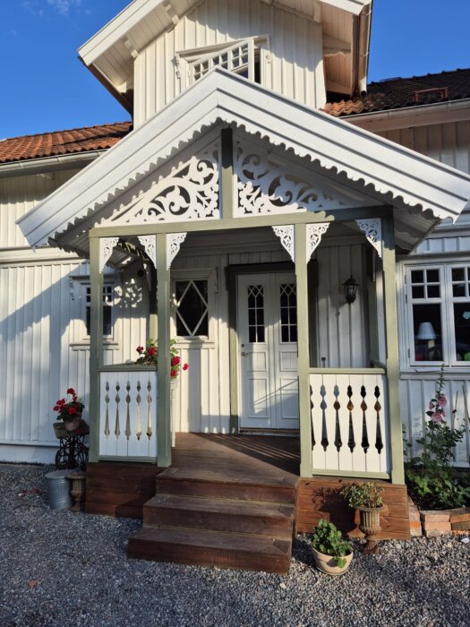 Vit träfarstukvist med snickarglädje och gröna detaljer. Röda blommor och krukor vid trappan. Huiset har en vit fasad.