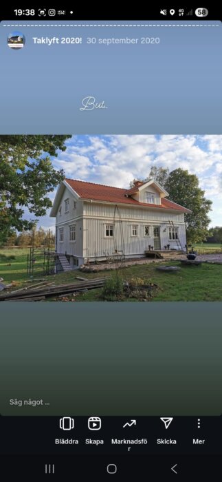 Vit husfasad med rött tak, omgiven av grönska och ett stort träd, med en nybyggd farstukvist och byggmaterial i förgrunden.