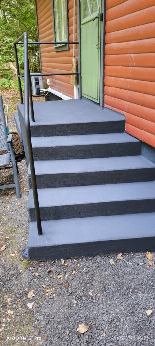 Nyrenoverad trappa i gråblå färg, liknande antracit, mot en röd fasad med en grön dörr. Trappans färg matchar husgrunden.