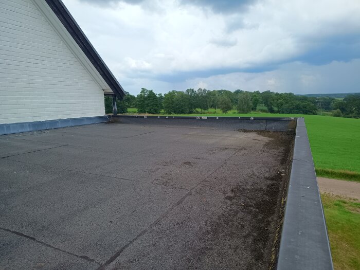 Platt tak med tjärpapp på en utbyggnad, omgiven av grönskande landskap med molnig himmel i bakgrunden.