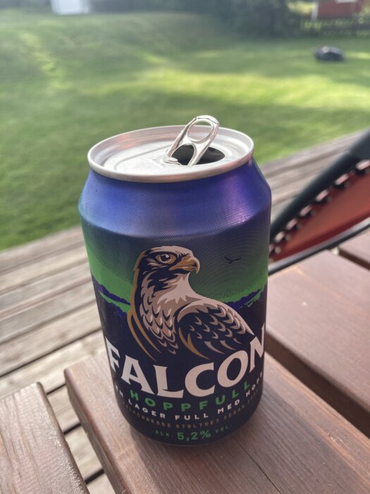 Öppnad burk med Falcon öl med design av en falk, placerad på ett träbord utomhus med gräs i bakgrunden.