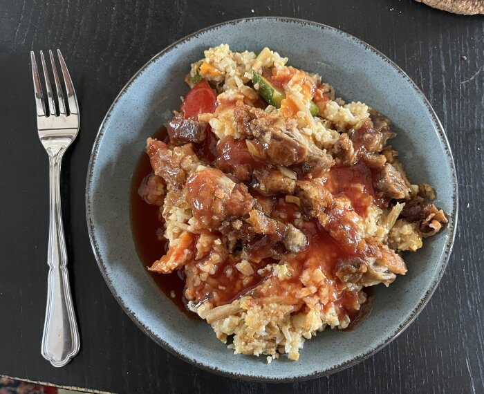 Stekt ris med grönsaker, anka och sötsur sås på en grå tallrik med en gaffel bredvid.