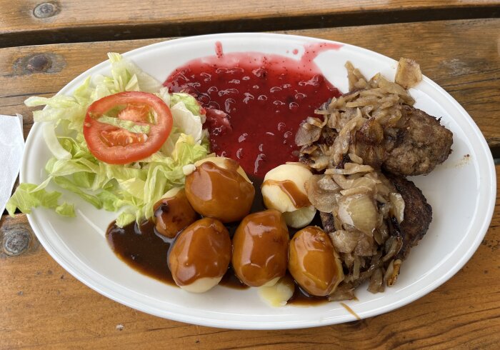 Tallrik med kålsallad, tomatskiva, pannbiff, stekt lök, kokt potatis med sås och lingonsylt på träbord.