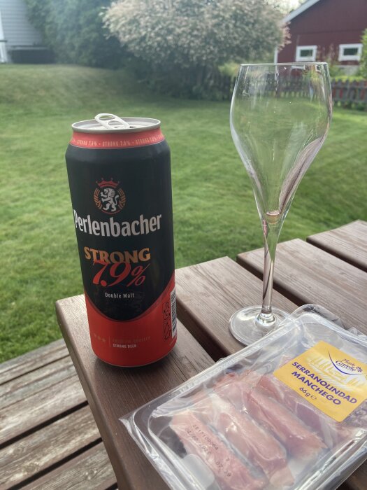 Burk med Perlensbacher Strong 7.9% öl, ett tomt vinglas och förpackad serranolindad manchego på ett trädäck.