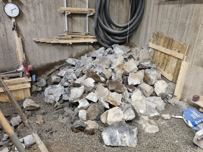 Hög av stenar i ett rum med betongväggar, hyllor och verktyg. Bland annat en borrmaskin och slangar syns på väggen.