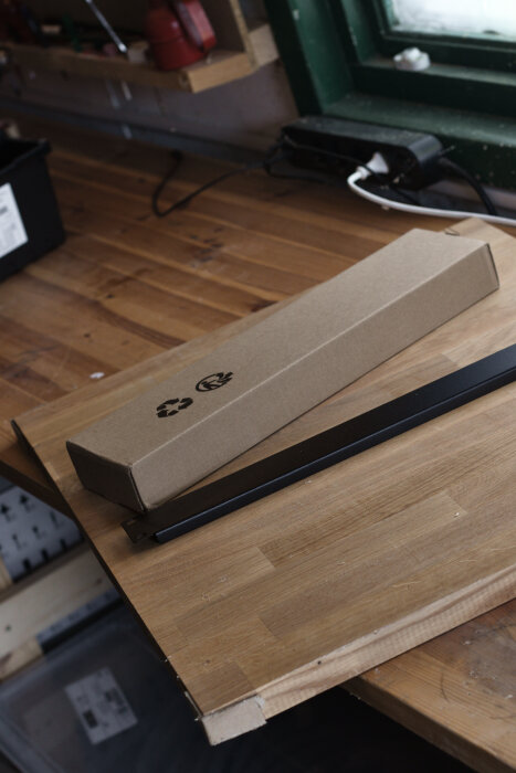 Kartong och svart metallstång på en arbetsbänk av trä, verkstadsverktyg synliga i bakgrunden.