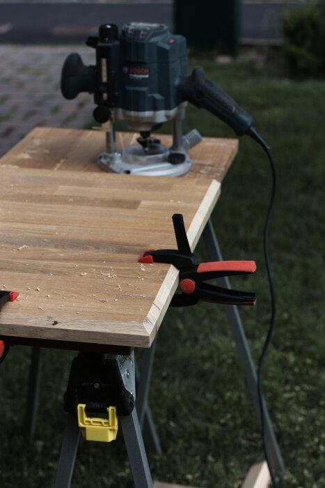 En ekskiva på ett arbetsbord med en fräs och tvingar, omgiven av gräs. Skivan har en bearbetad kant och spånor syns på ytan.