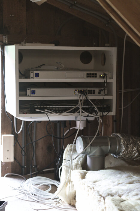 Serverrack monterat på vinden med nätverksutrustning och synliga sladdar. Träram och isoleringsmaterial syns i förgrunden.