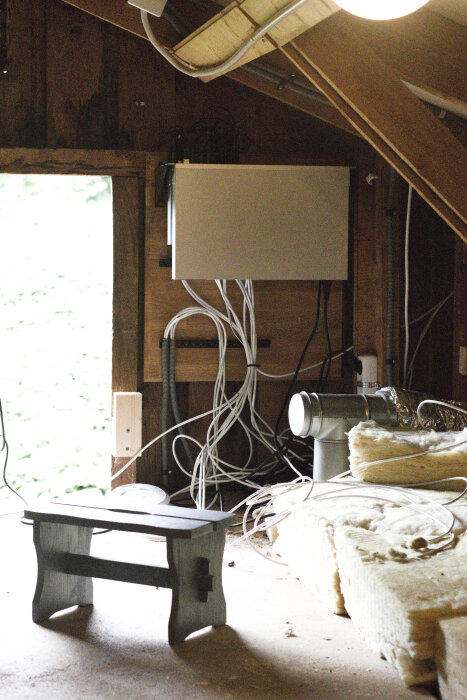 Serverrack monterat på vägg med sladdar hängande i en arbetsmiljö på en vind. En pall står på golvet och det finns rörisolering.