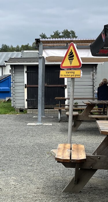 Varningsskylt för glass vid träbord utomhus, i närheten av en byggnad med svarta dörrar och metalltak.