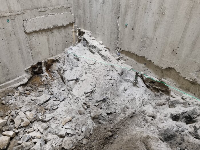 Betongyta med löst material och synliga kilar vid en vägg, där delar har flagnat och berg är synligt.