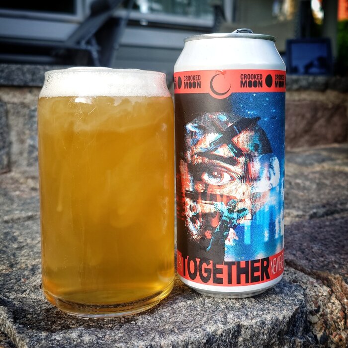 En glas öl bredvid en Crooked Moon burk med konstnärlig etikett, står på stenläggning utomhus.