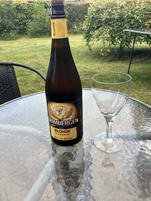 Flaska Grimbergen Blonde och vinglas på ett trädgårdsbord i en grönskande trädgård.