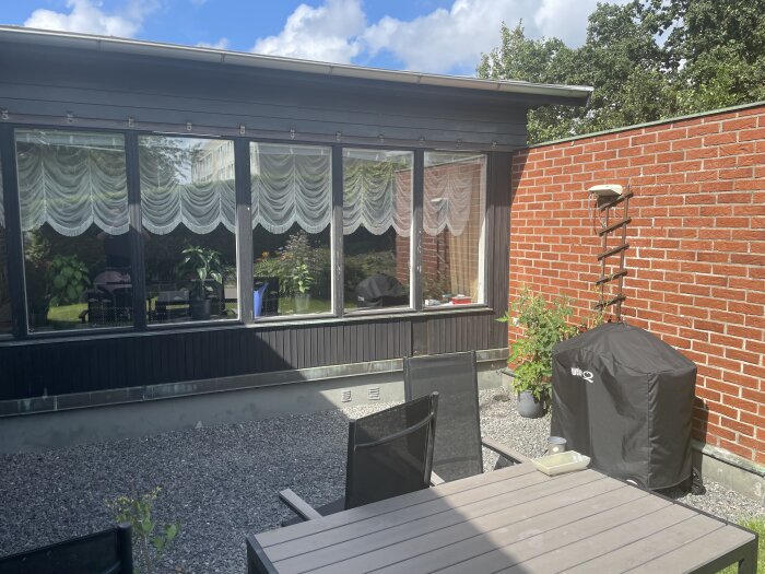 Utomhusbild av en altan med svart panel och mörkblå överdel vid fönstren, samt en grill med skydd, trädgårdsbord och stolar i förgrunden.
