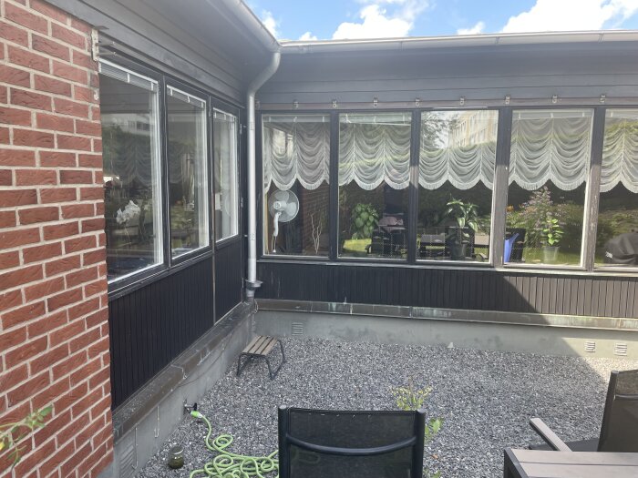 Svart panel vid altan med mörkblå färg ovanför fönstren, vita gardiner och blommor i fönstren. Gårdsplan med grus och en grön vattenslang.