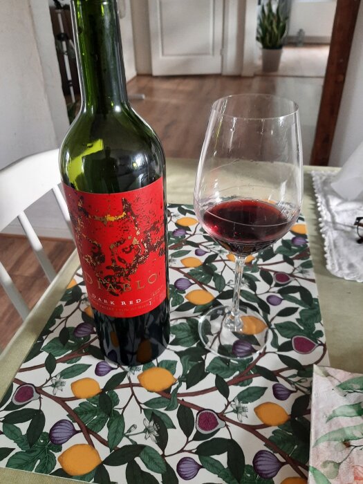 Rödvinsflaska och glas med rödvin på ett mönstrat bord.