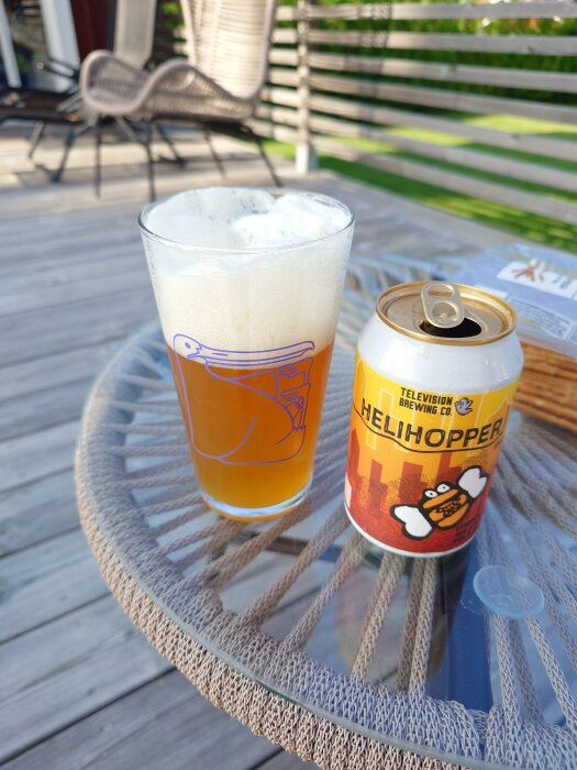 Ölglas med skummande öl och en öppnad burk med texten "Helihopper" på ett utebord av rotting.