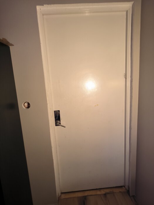 Vit dörr med enkel dörrkarm, ljus vägg och trägolv. Dörrhandtag i metall, omgivningen inbjuder till förslag på förbättringar runt karmen.