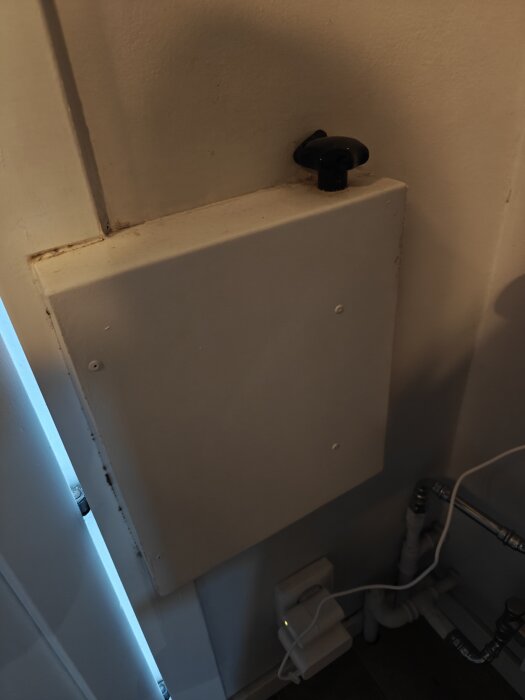 Vit ventilationslåda med svart reglage på en vägg, delvis dold av dörr. Rör och elektriska ledningar syns nedanför lådan.