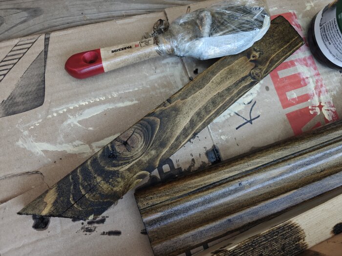 Bitar av trä med olika pigmentering, en pensel med rött handtag, och en plastinpackad pensel. Bakgrund av kartong med text och färgrester.