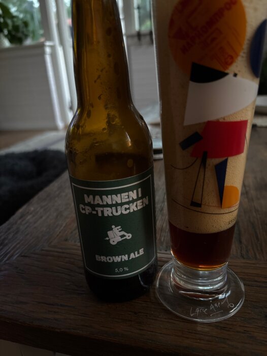 Brun glasflaska med etiketten "Mannen i CP-trucken" och ett glas öl på ett mörkt träbord i ett ljust rum.
