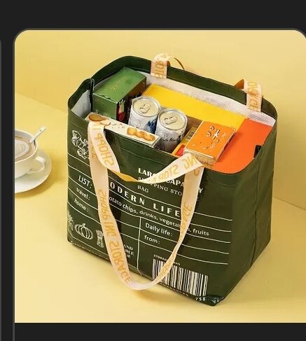 En stor grön shoppingkasse fylld med varor som burkar och kartonger, med beige handtag, står på ett bord.