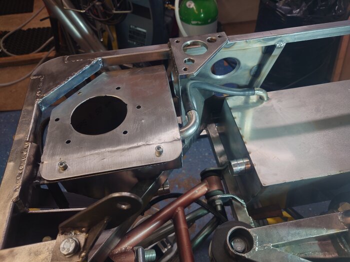 Metallkonstruktion med svetsade komponenter och hål för avluftning vid en tank, omgiven av verktyg och rör.