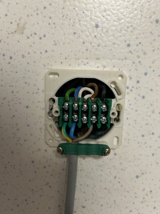 Äldre fast inkopplad elinstallation med synliga kablar och skruvanslutningar mot en vit, stänkmönstrad vägg.