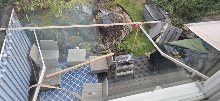 Vy över en uteplats med rottingmöbler, en blå matta och diverse byggutrustning, inklusive en stege och en bräda. Bakom syns en jordhög och buskar.