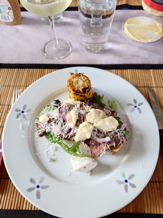 Tallrik med tunnt skuren rostbiff på salladsbädd, rostad levain, parmesansås, tryffelaioli, riven parmesan, och majskolv. Vin och vattenglas i bakgrunden.